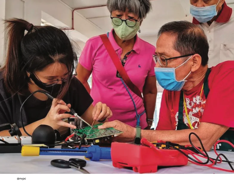 A young woman solders an electronic circuit board as volunteers look on, at a Repair Kopitiam repair workshop in Singapore