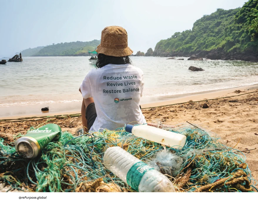 Plastic waste and fishing nets on a beach, facing a person wearing a rePurpose t-shirt reading “Reduce Waste, Revive Lives, Restore Balance”