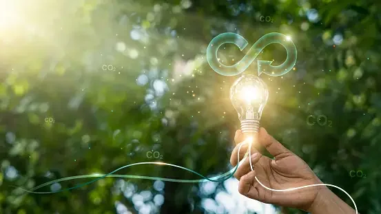 A hand holding a lit light bulb topped with an infinity symbol, against a background of foliage